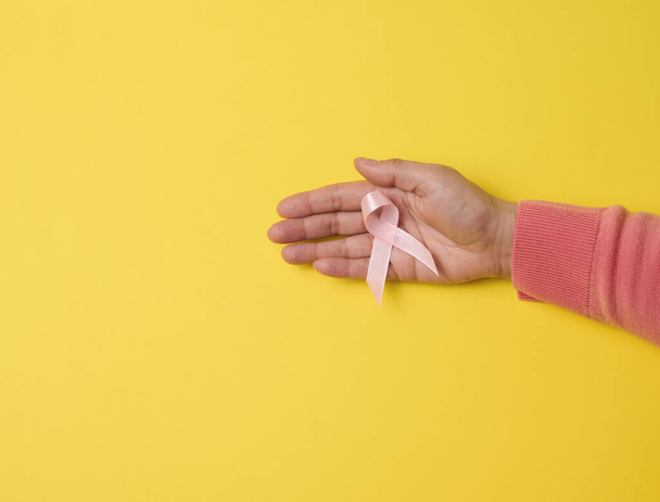 female hand hold a pink silk ribbon in the form of a loop on a yellow background. Symbol of the fight against breast cancer - Photo, image