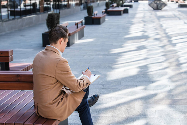 Happy man in beige coat and glasses sits on bench near modern business center, getting ready to draw sketch, writing text - Foto, Imagem