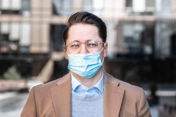 Serious business man in glasses and mask on street, against background of modern business center, covid-19 pandemic, concept of protected business. - Fotoğraf, Görsel