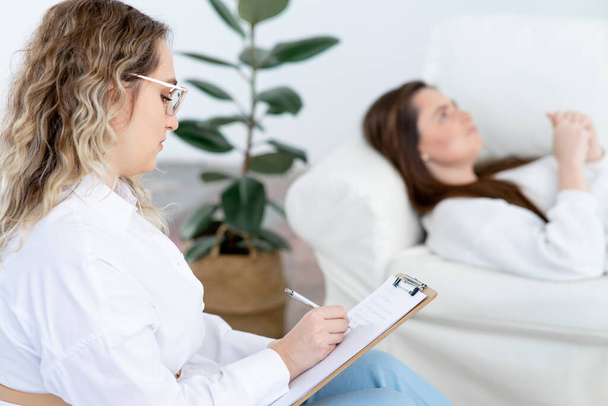 Concentrated blonde woman psychologist with overweight listens to lady patient making notes on clipboard in clinic office side view - Foto, imagen