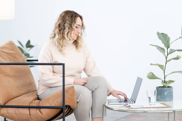 Blonde woman with overweight in casual clothes works on laptop computer sitting in armchair at small table in office side view - Foto, immagini