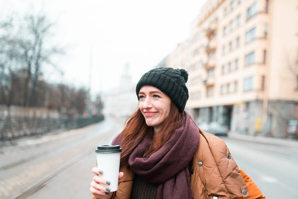 A woman walks around the city with a cup of coffee, warming drink in the cold season. Warm winter hat, scarf and winter jacket. Blurry image of the city. - Foto, Imagem