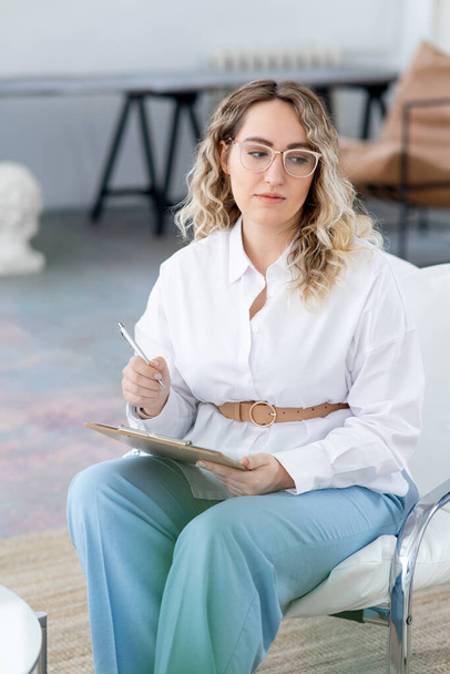 A blonde doctor woman is sitting in a chair with a clipboard and a pen. Professional help of a psychotherapist. Bright office. Mental health. White shirt and blue trousers. - Fotoğraf, Görsel
