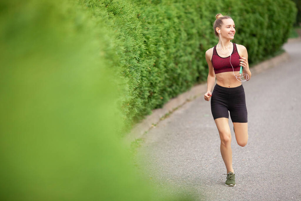 Morning jog in park area. Jogging with music on your phone. Music in headphones. Running woman in full growth runs us. Dressed in burgundy top and black shorts. Blurred green branches in foreground. - Foto, Imagen