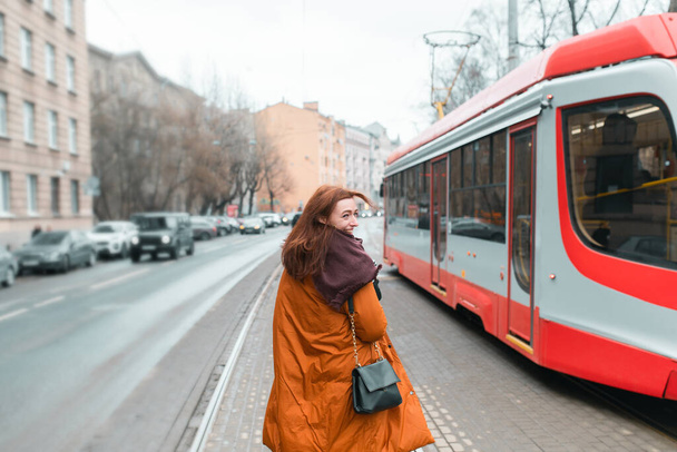 A woman goes to public transport. Modern public transport. City street. Long warm coat. Happy young woman. Woman getting on the tram. A trip to work. - Photo, Image