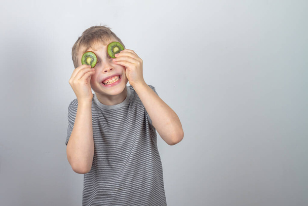 A cheerful Caucasian boy closed his eyes with slices of green kiwi fruit on a gray background in the studio. - Foto, imagen