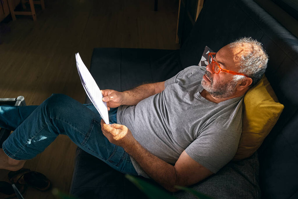 ソファに座って読書眼鏡をかけたハンサムな熟年が家で仕事をしている間に書類を見ている。リモートワークの概念. - 写真・画像