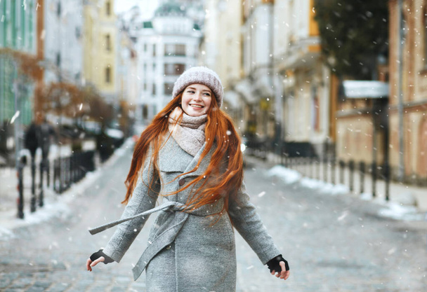 Позитивная рыжая длинноволосая женщина носит стильное пальто, гуляя по улице во время рождественских праздников со снегом. Пространство для текста - Фото, изображение