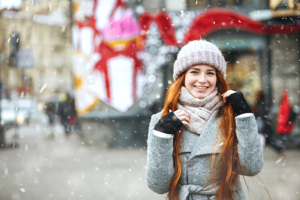 Joyful red haired woman wears coat walking at the city during snowfall. Space for text - Photo, Image