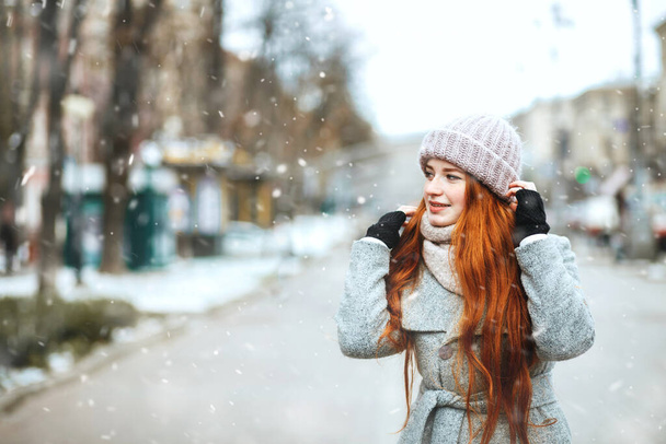 Emocjonalnie ruda kobieta nosi płaszcz spacerując po mieście podczas opadów śniegu. Miejsce na tekst - Zdjęcie, obraz