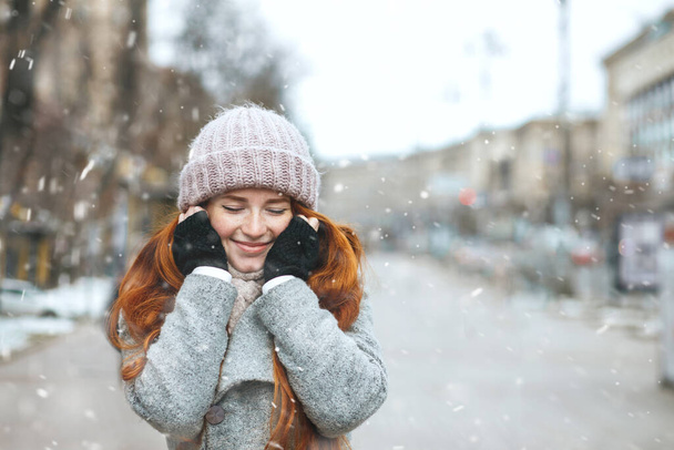 Delightful red haired woman wears coat walking at the city during snowfall. Space for text - Photo, Image
