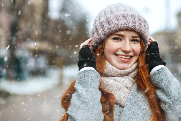 Fashionable red haired woman wears coat walking at the city during snowfall. Space for text - Photo, Image