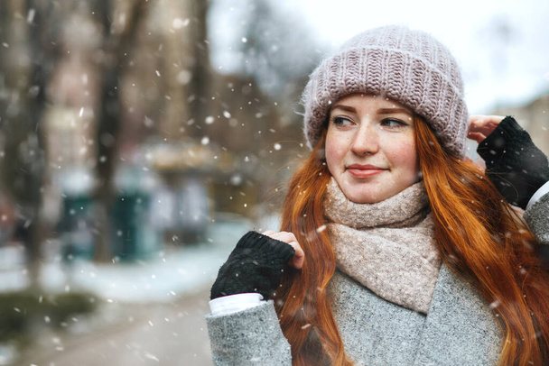 Wspaniała rudowłosa kobieta nosi płaszcz spacerując po mieście podczas opadów śniegu. Miejsce na tekst - Zdjęcie, obraz