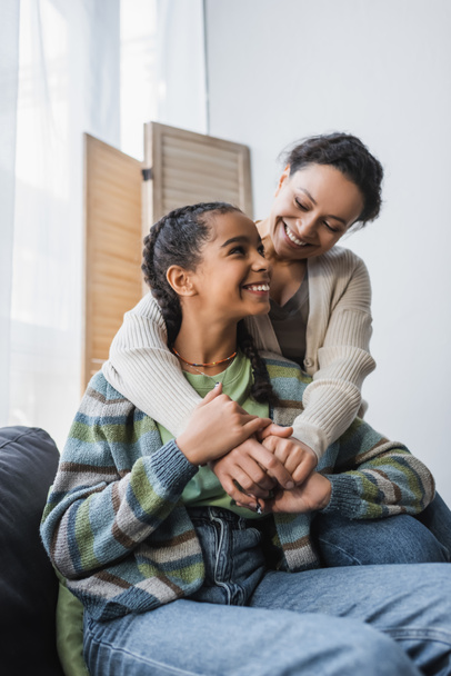 happy african american woman embracing teenage daughter sitting on couch at home - Photo, image