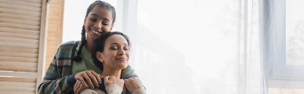 teenage african american girl hugging happy mom with closed eyes near window at home, banner - Photo, Image
