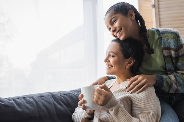 cheerful african american teenage girl hugging happy mother sitting on couch with tea cup - Photo, Image