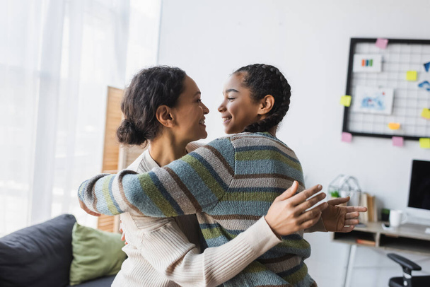 joyful african american mother with teen daughter looking at each other while embracing at home - Photo, image