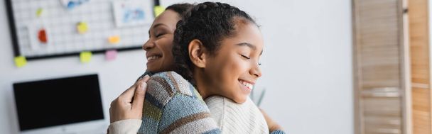 vreugdevolle Afrikaanse Amerikaanse vrouw en tienermeisje omarmen met gesloten ogen thuis, banner - Foto, afbeelding
