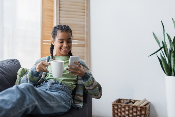 smiling african american teenage girl chatting on mobile phone while holding cup of tea - Photo, Image