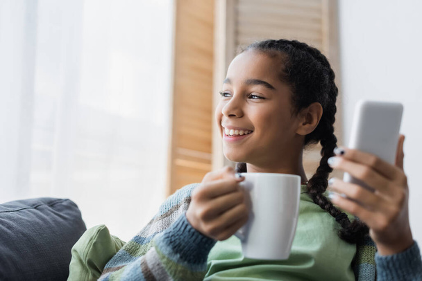 heureux afro-américain adolescent fille tenant tasse de thé et smartphone flou à la maison - Photo, image