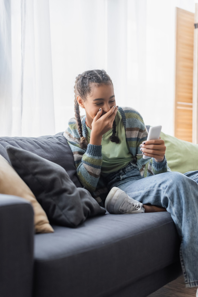 african american girl laughing and covering mouth with hand while sitting on couch and looking at smartphone - Photo, Image