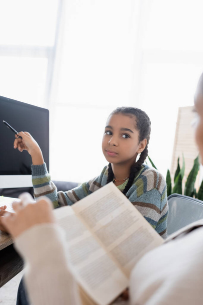 Africano americano adolescente menina apontando com caneta no monitor com tela em branco perto mãe turva com livro - Foto, Imagem