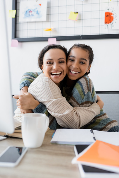 happy african american mother and daughter embracing near blurred computer at home - Photo, Image