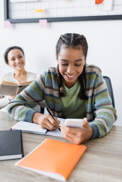 gelukkig Afrikaans amerikaans tiener meisje schrijven in notebook in de buurt van moeder met boek glimlachen op wazig achtergrond - Foto, afbeelding