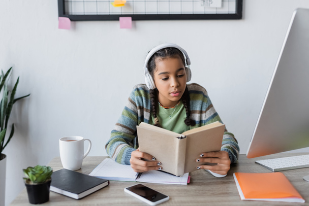 african american teenage girl in headphones reading book near computer and smartphone with blank screen - Photo, Image