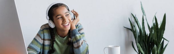 gelukkig Afrikaans amerikaans meisje in hoofdtelefoon glimlachen in de buurt computer monitor thuis, banner - Foto, afbeelding
