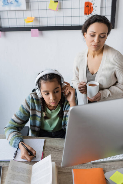 Kulaklıklı Afro-Amerikan kız bilgisayarın yanında not defterine çay fincanı ile anne yazıyor. - Fotoğraf, Görsel