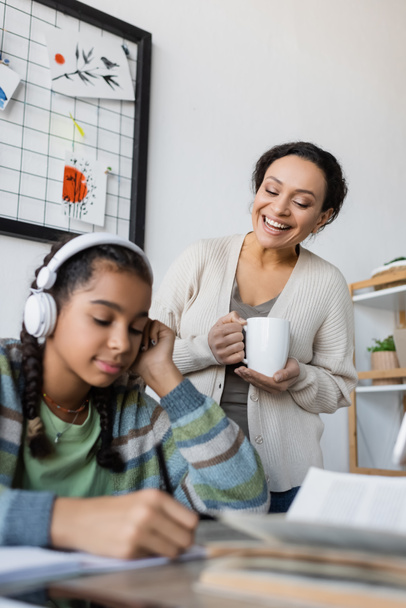 african american girl in headphones doing homework near smiling mother with cup of tea - Photo, Image