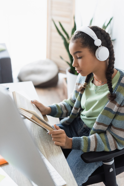 african american schoolgirl in headphones reading book near blurred computer monitor - Foto, Imagen