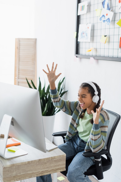 excited african american teen girl in headphones showing wow gesture near computer monitor - Photo, Image