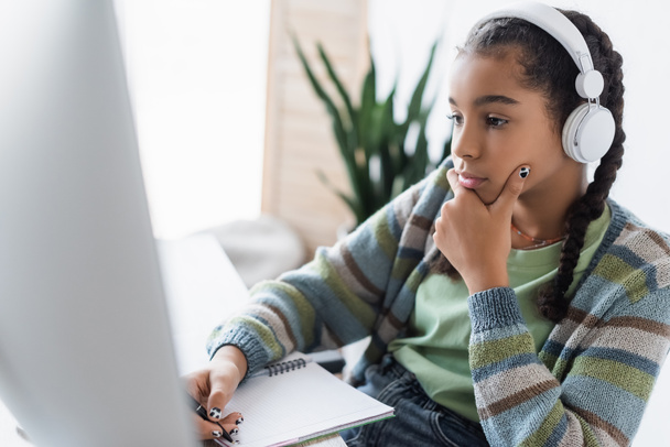 přemýšlivý africký americký školačka ve sluchátkách psaní v notebooku v blízkosti rozmazané monitor počítače - Fotografie, Obrázek