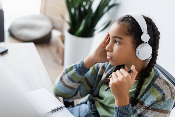 reflexivo afroamericano adolescente chica en auriculares celebración de la pluma y mirando hacia otro lado mientras estudia en casa  - Foto, Imagen