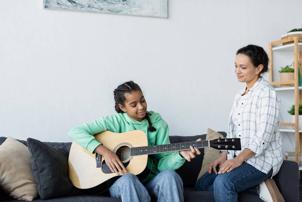 Усмішка африканського американця, який сидить на дивані біля підліткової дочки, граючи на гітарі. - Фото, зображення