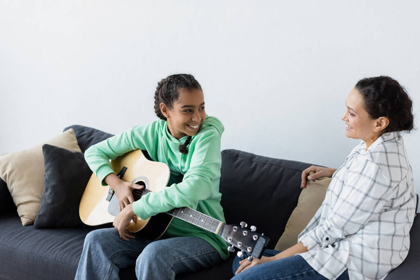 happy african american teen girl with acoustic guitar sitting on sofa near smiling mom - Fotó, kép
