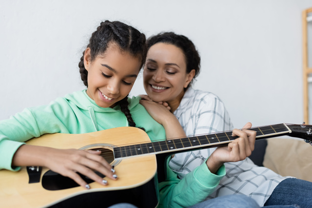 happy african american woman smiling near teenage daughter playing guitar at home - Fotó, kép