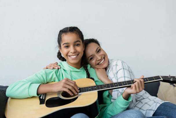 щаслива афроамериканка, яка обіймає усміхнену дочку, граючи на гітарі вдома
 - Фото, зображення