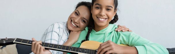 joyful african american woman hugging teenage daughter playing guitar, banner - 写真・画像