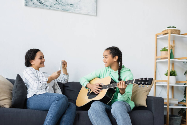 Африканська американка з смартфоном фотографує доньку, яка грає на гітарі вдома. - Фото, зображення