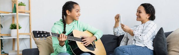 Африканська американка з мобільним телефоном робить фото усміхненої дочки, яка грає на гітарі вдома, банер. - Фото, зображення