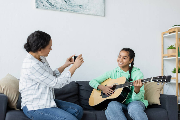 joyful african american teenage girl playing guitar near mom taking photo on cellphone - 写真・画像