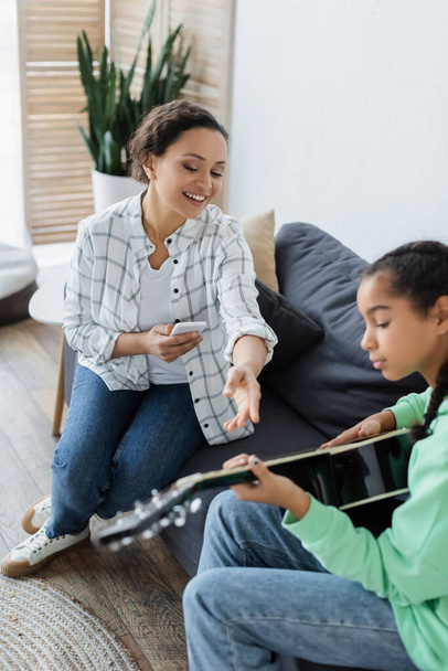 african american woman with smartphone pointing with hand near daughter playing acoustic guitar - Photo, Image