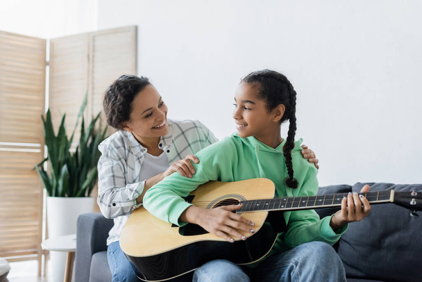 uśmiechnięta afrykańska dziewczyna patrzy na szczęśliwą mamę podczas gry na gitarze w domu - Zdjęcie, obraz