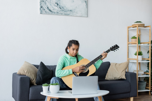 teenage african american girl learning to play guitar near laptop while sitting on sofa with pillows  - Fotoğraf, Görsel
