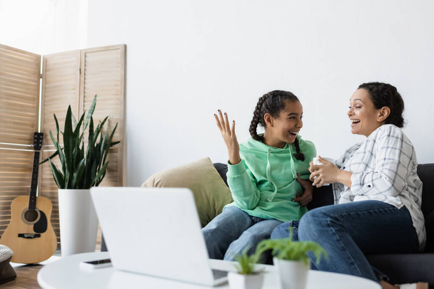 excited african american girl gesturing near mom while watching film on blurred laptop - 写真・画像