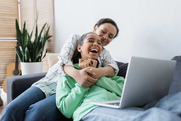 african american woman hugging laughing daughter while watching film on laptop - Foto, immagini