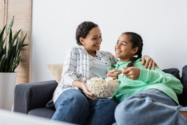 happy african american woman with bowl of popcorn sitting on sofa with teenage daughter - Фото, изображение